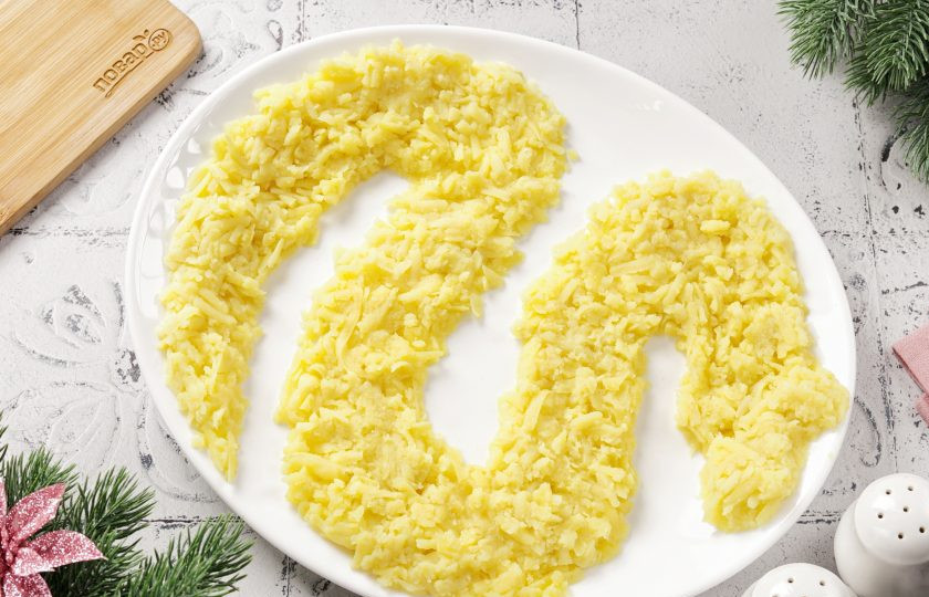 1. Этап. Селедка под шубой на год Змеи 2025: Картофель натрите на крупной терке и выложите первым слоем в виде символа года — змеи. Картофель посолите и смажьте майонезом.