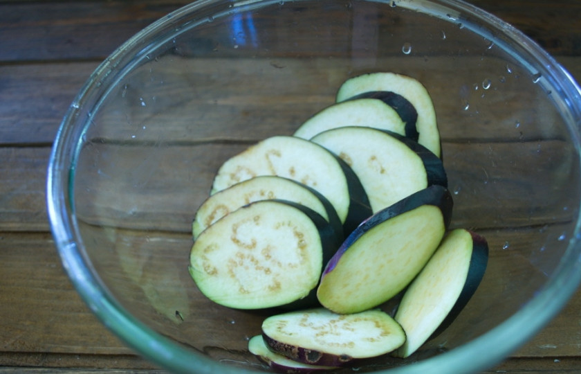 1. Этап. Баклажаны, запеченные с помидорами и сыром: Баклажаны нарезаем колечками. Кладем в миску. Солим (1 ч. ложка соли) и вливаем 1 стакан воды. Оставляем на 30 минут. Этот шаг можно и пропустить, если используете свежие молодые овощи.
