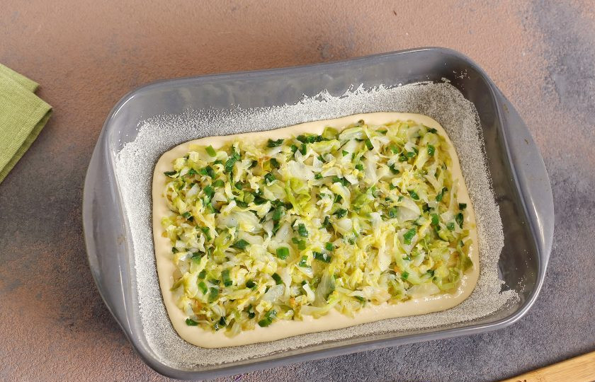 1. Этап. Капустный пирог, который тает во рту: Форму для запекания смажьте маслом. Дно и бока обсыпьте мукой или манкой. Вылейте половину порции теста, а сверху равномерно распределите всю начинку.