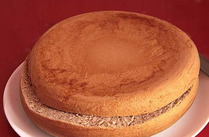 1. Этап. Торт с вишней и орехами: Как только бисквит остынет, начните формировать торт. Разрежьте выпечку вдоль так, чтобы нижняя часть была больше (2 см и 1,5 см). Большую сторону нарежьте на куски по 2,5 см, а меньшую уложите на тарелку для подачи.