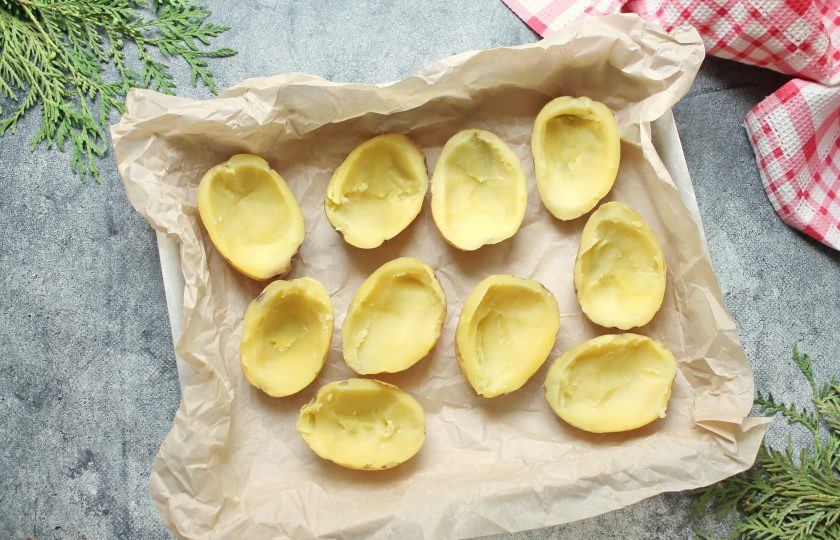 1. Этап. Запеченный картофель с беконом и сыром: Теплый картофель разверните и разрежьте на две части вдоль. Ложкой удалите мякоть из каждой половинки, оставив стенки толщиной 5-6 мм. Выложите картофель на противень застеленный пергаментом срезом вверх, немного посолите.