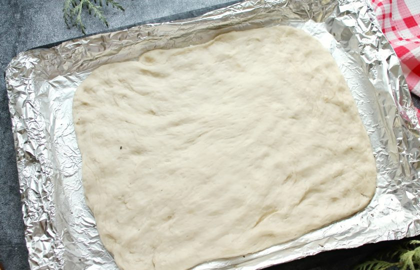 1. Этап. Фокачча с пармезаном и розмарином: Противень застелите пергаментом или фольгой и смажьте растительным маслом. Переложите подошедшее тесто в противень и руками распределите в лепешку прямоугольной формы. Накройте фокаччу пленкой и оставьте в тепле на 30 минут.