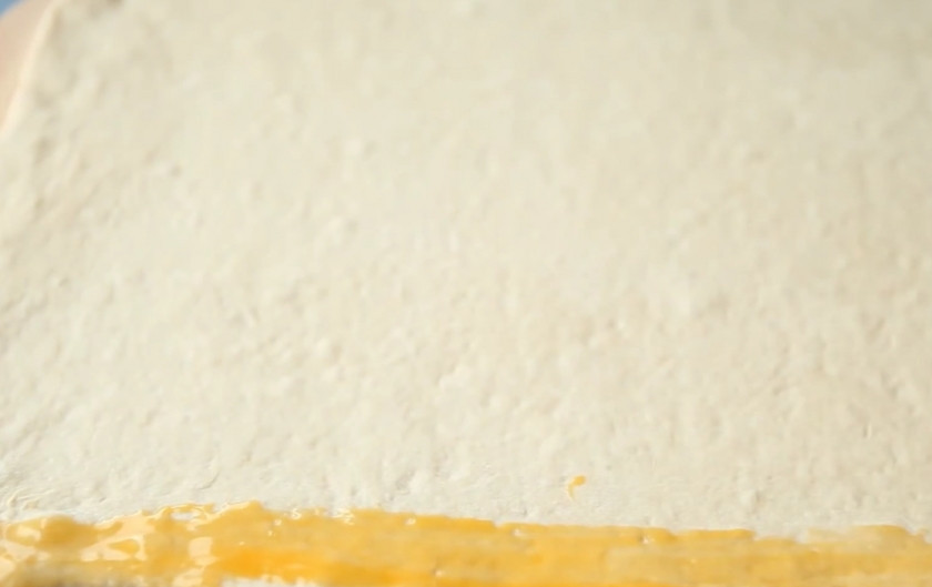 1. Этап. Миндальные бантики (слойки): 3. Возьмите дрожжевое слоеное тесто (у меня оно домашнего приготовления) и раскатайте в пластину размером примерно 110 на 36 сантиметров. Смажьте один край теста яичным желтком.