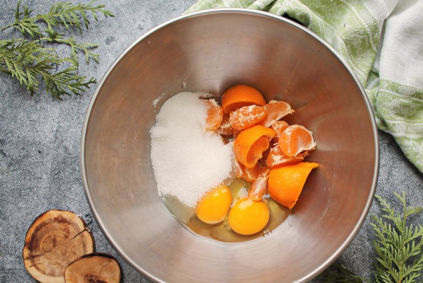 1. Этап. Кекс Мандариновая песня: Один мандарин почистите и разберите на дольки. Второй мандарин порежьте на кусочки вместе с кожурой и выложите в миску. Всыпьте к мандаринам сахар и разбейте яйца.