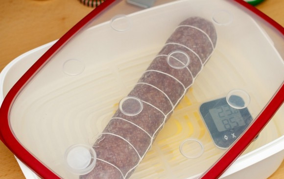 1. Этап. Итальянская домашняя салями: На дно ёмкости налейте воды. В неё уложите сетку, а сверху положите салями, чтобы она погрузилась в воду. Закройте всё крышкой. Оставьте колбасу в таком виде на 3 дня.