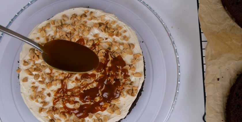 1. Этап. Торт Сникерс с соленой карамелью: 9. У коржей срежьте верхушки. Первый корж смажьте кремом, посыпьте рубленым арахисом и распределите карамельную начинку. Повторите с остальными коржами. Верхних корж и бока смажьте кремом, украсьте кремом, арахисом и карамельной начинкой.