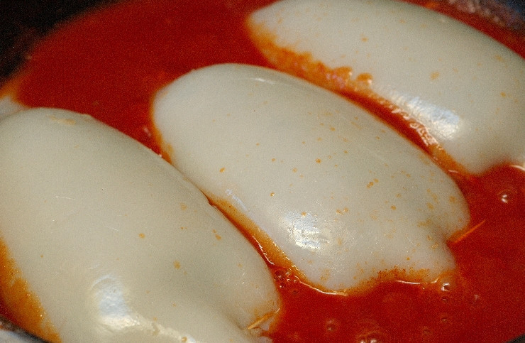 1. Этап. Кальмары, фаршированные курицей и грибами: Далее мы берем толстостенную кастрюлю и наливаем в нее растительное масло, ставим кастрюлю на огонь. В разогретое масло выкладываем нарезанную половинку луковицы и жарим лук до прозрачности, затем добавляем измельченный чеснок и лавровый лист, жарим все еще пол минутки. После этого вливаем в кастрюлю вино и ждем, пока оно почти полностью выпарится. Теперь добавляем томатную пасту и бульон, солим и приправляем все хлопьями красного перца по вкусу. Как только соус закипит, выкладываем в него кальмары.