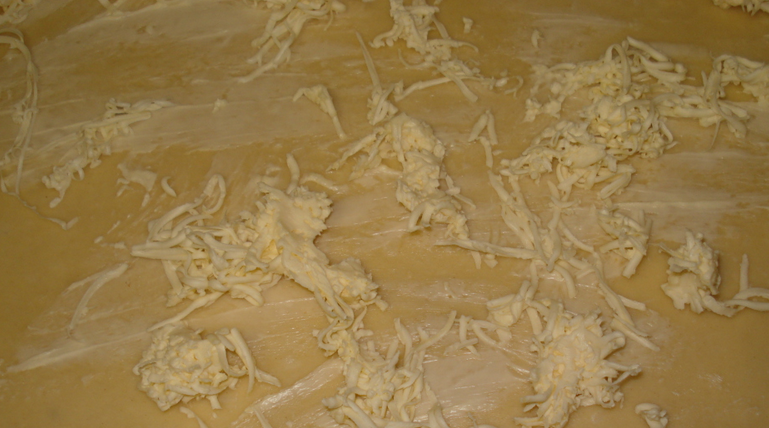 1. Этап. Булочки из слоеного дрожжевого теста: Раскатайте тесто в квадратный пласт. Сверху натрите на терке замороженный маргарин.