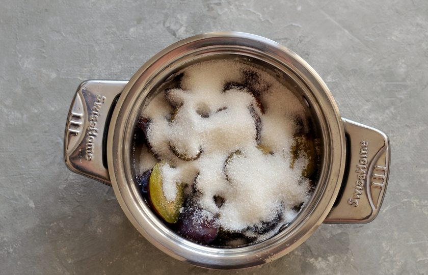 1. Этап. Варенье с орехами: Переложите сливы в кастрюлю с толстым дном, затем насыпьте сахар. Налейте воду, капните ванильный сироп и доведите до кипения на среднем огне.