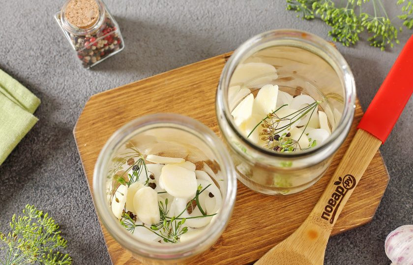 1. Этап. Маринованные помидоры с луком и чесноком на зиму: На дно чистых стерилизованных банок выложите нарезанный пластинами чеснок, лук нарезанный кольцами, зонтики укропа и перец горошком.