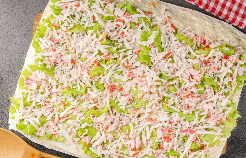 1. Этап. Рулет из лаваша с крабовыми палочками: Накройте начинку вторым лавашом и так же смажьте майонезом с чесноком. Выложите листья салата и тёртые на крупной тёрке крабовые палочки.