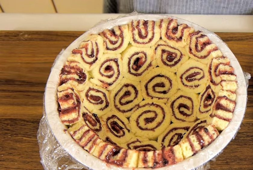 1. Этап. Торт Шарлотта: 4. Желатин залейте холодной кипяченой водой, перемешайте и оставьте для набухания. Миску для торта выстелите пищевой пленкой. Выложите поверхность миски рулетом, нарезанным на кружочки толщиной 1,5-2 см, плотно прижимая кусочки друг к другу.