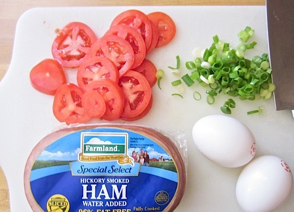 1. Этап. Пицца к завтраку: 4. Помойте и нарежьте помидор кольцами, промойте зеленый лук и порежьте. Нарежьте тонкими кусочками ветчину, натрите сыр на средней терке.