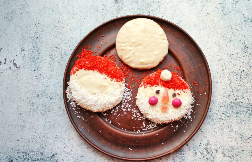 1. Этап. Бисквитное печенье Дед Мороз: Белый шоколад растопите на водяной бане до однородности, так чтобы вода не касалась чаши с шоколадом. Нанесите при помощи кондитерской кисточки на каждую печеньку. Сверху сделайте шапочку из красной кокосовой стружки и бороду с усами из белой, установите носик и рот из мармелада. Из шоколадных капель сделайте глазки, а из красного драже щеки. Заготовку из маршмеллоу сначала опустите одной стороной среза в шоколад, затем в белую кокосовую стружку и установите ее на шапке. При необходимости окуните второй срез также в шоколад.