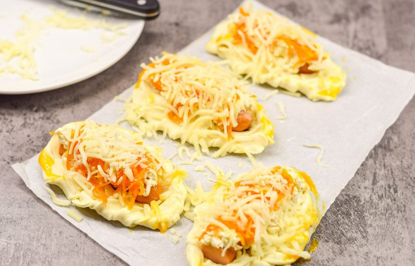 1. Этап. Плетеные Хот-доги из слоеного теста с плавленым сыром: Поверх корейской моркови выложите тертый сыр. Края заготовок смажьте взбитым желтком. Переложите заготовки на противень, отправьте в духовку.