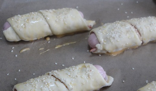 1. Этап. Сосиски в пикантной обертке: Обернуть сыром внутрь вокруг сосиски. Уложить на противень, выложенный пергаментом. Смазать взбитым яйцом и посыпать кунжутом.