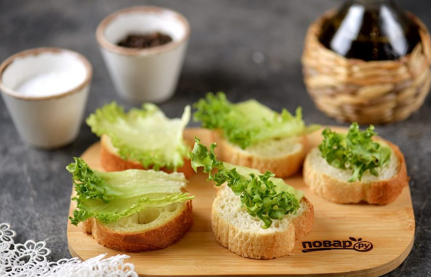 1. Этап. Канапе с курицей: Выложите багет на разделочную доску, на каждый ломтик хлеба выложите листик латука. Если планируете подавать канапе сразу же после приготовления, тогда можете хлеб слегка смазать любимым соусом.