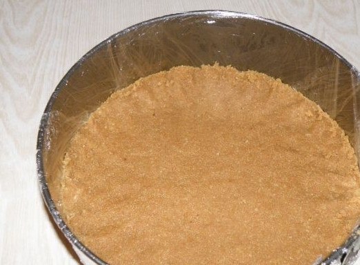 1. Этап. Чизкейк с киви без выпечки: Печенье измельчаем в крошку. Масло необходимо растопить на огне, после остудить и соединить с печеньем. Формируем тесто и выкладываем его ровненьким слоем в застеленную пленкой форму для запекания. Отправляем в холодильник.