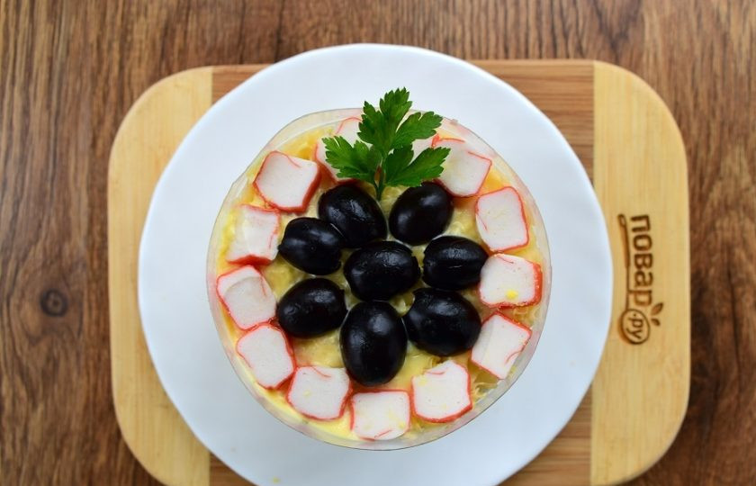 1. Этап. Крабовый салат с черносливом: Верх салата украсьте по своему вкусу. Я по краю положила ломтики крабовых палочек, в центре — половинки маслин без косточек и зелень.