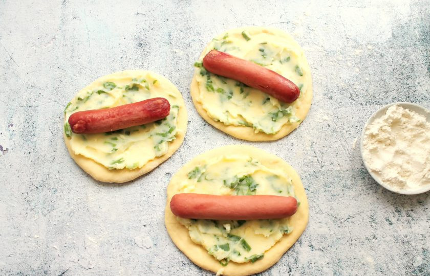 1. Этап. Пирожки Два в одном: Тесто разделите на три части и раскатайте в лепешки по длине сосисок. Смажьте их поверхность картофельным пюре с луком. На середину выложите по одной сосиске.