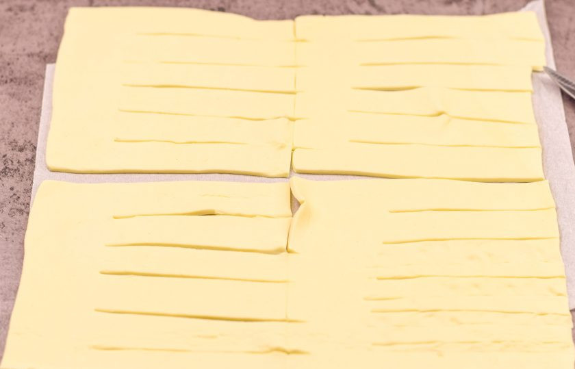 1. Этап. Плетеные Хот-доги из слоеного теста с плавленым сыром: Слоеное тесто раскатайте, разрежьте на 4 прямоугольника. Каждую заготовку разрежьте на 6 полосок, но не доходя до самого конца.