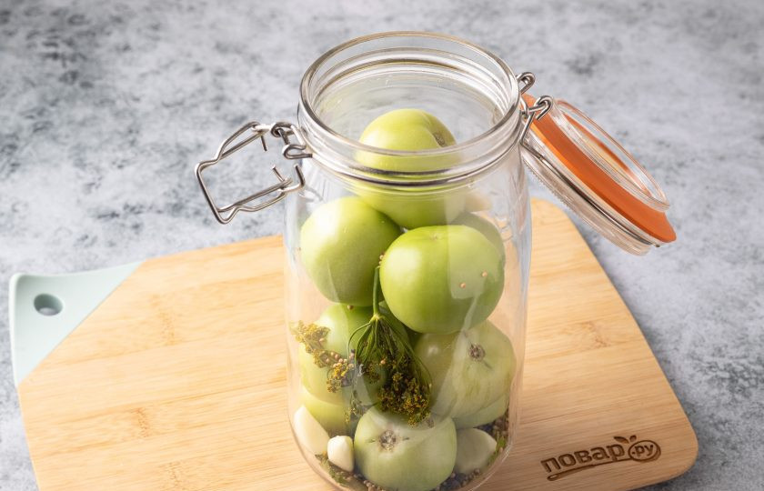 1. Этап. Соленые зеленые помидоры: Заполните банку зелеными помидорами. В свободные места добавьте оставшийся чеснок, зонтики укропа, гвоздику, семена кориандра и горчицы.