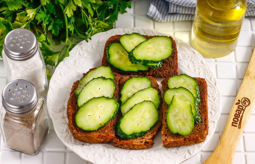 1. Этап. Тосты с селедкой: Промойте огурцы в воде, удаляя шипы и пыль, срежьте хвостики с овощей. Нарежьте их слайсами и выложите на тосты.