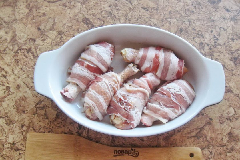 1. Этап. Куриные ножки в беконе в духовке: Форму смажьте подсолнечным маслом и выложите голени в беконе. Накройте фольгой и отправьте в духовку, разогретую до 185-190 градусов. В форму можно налить немного воды или бульона.