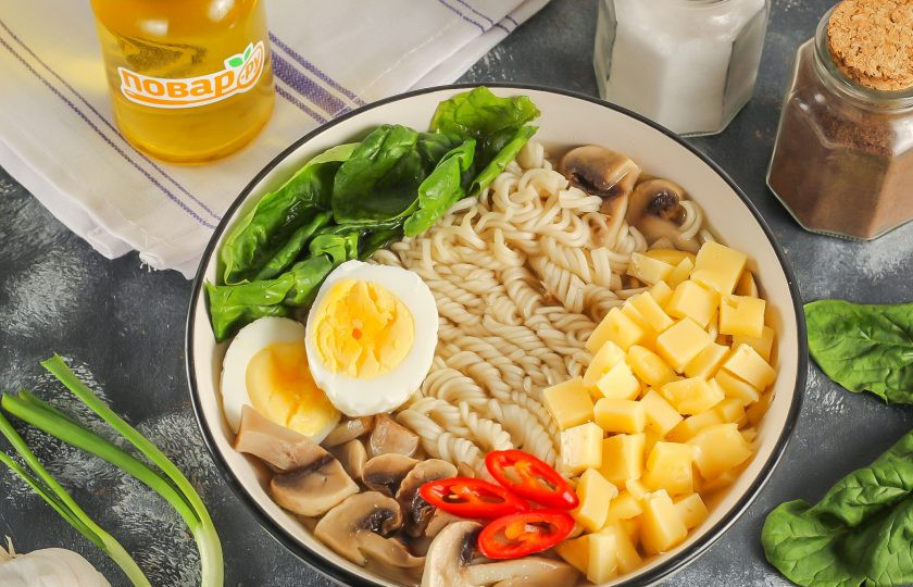 1. Этап. Рамен с сыром: Очистите отварное куриное яйцо, промойте его в воде и разрежьте пополам. Нарежьте сыр средними или мелкими кубиками и добавьте в емкость вместе с яйцом. Аккуратно нарежьте острый перец и выложите нарезку на лапшу.