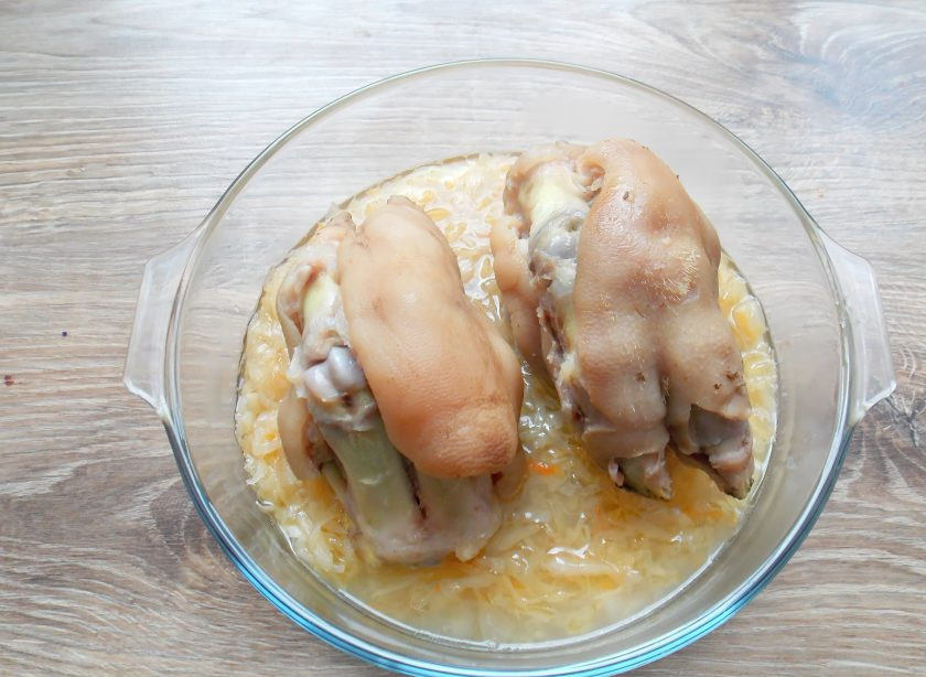 1. Этап. Свиные ножки с капустой: В форму для запекания переложите капусту и сверху выложите свиные ножки. Добавьте 2-3 столовые ложки бульона.