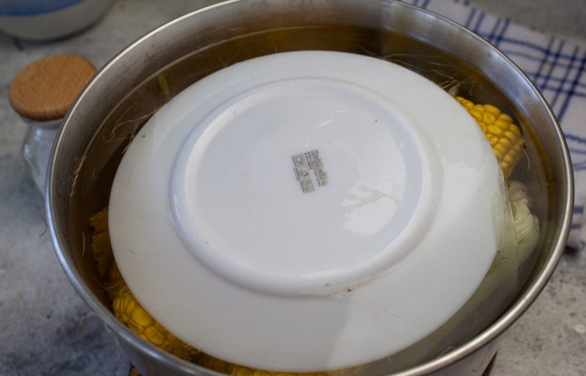 1. Этап. Кукуруза в початках: Для того, чтобы кукуруза не поднималась на поверхность, выложите сверху тарелку меньшего размера. Доведите до кипения, убавьте нагрев до минимального. Накройте кастрюлю крышкой и варите молодую кукурузу максимум 15 минут. Затем отключите нагрев и оставьте еще минут на 15 кукурузу в горячей воде.