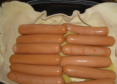 1. Этап. Пирог с сосиской и картошкой: Разделите тесто на 2 равных части, каждую из них раскатайте в пласт. Один пласт помещаем на дно формы для выпечки, выкладываем на него картофель, порезанный тонкими ломтиками, и сосиски, разрезанные вдоль пополам.