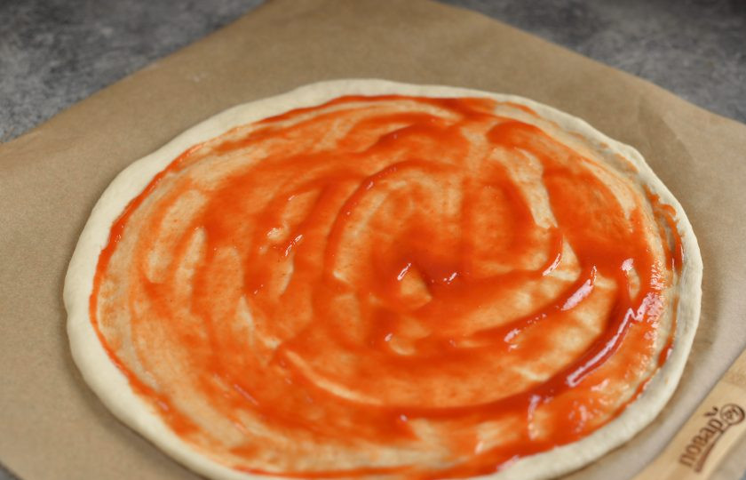 1. Этап. Пицца Каприччио: Пергамент присыпьте мукой, выложите на него тесто, раскатайте тонким кругом, смажьте томатным соусом любимого производителя.