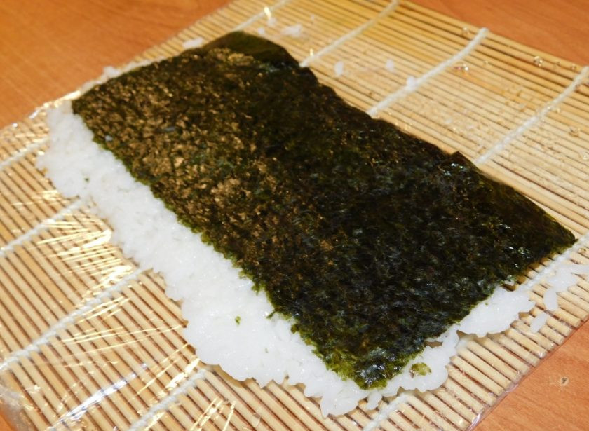 1. Этап. Роллы Бонито: Половину листа нори уложите на циновку блестящей стороной вниз. Распределите по нори тонкий слой риса так, чтобы с одной стороны осталось около 1 см. нори, а с другой стороны рис выступал тоже примерно на 1 см. Затем переверните, чтобы рис оказался внизу.