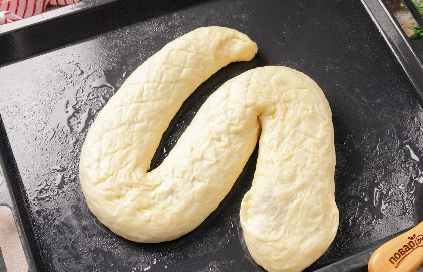 1. Этап. Новогодний пирог Змейка: Переверните рулет швом вниз и слегка потяните руками, выложите на смазанный маслом противень и сформируйте тело и голову. Можно попробовать сделать насечки ножом, для имитации кожи. Накройте заготовку полотенцем и дайте полежать 30 минут.