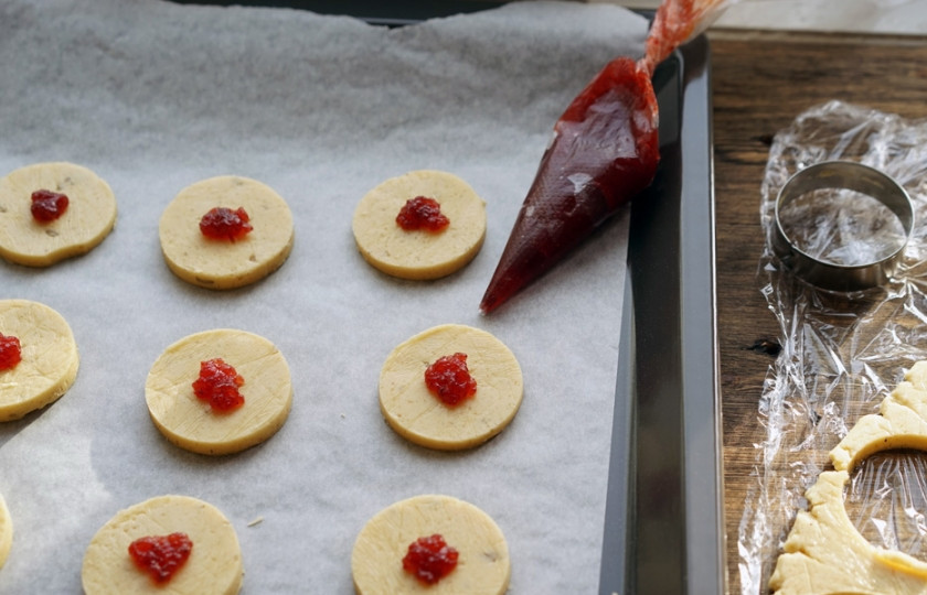 1. Этап. Песочное печенье с джемом: Раскатайте тесто. С помощью стакана вырежьте из него круги (это и будет наше печенье). При помощи кондитерского мешка или пакета выдавите немного джема на каждый кружок. Выложите будущие печенья на противень, предварительно застеленный бумагой для запекания.