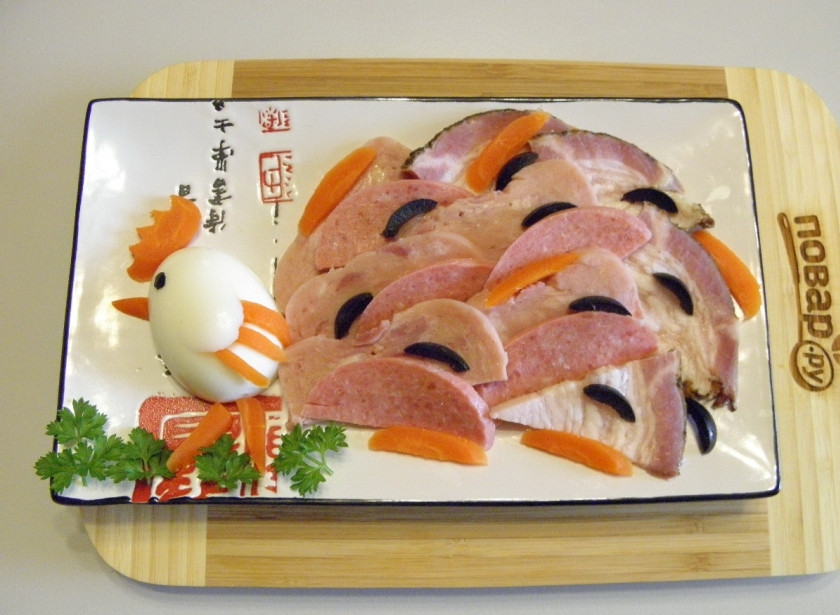 1. Этап. Мясная нарезка Хвост петуха: Возьмите тарелочку прямоугольной формы, так удобнее будет расположить петушка с хвостом. Яйцо разрежьте на две половинки. Разложите по тарелочкам, из оливок сделайте глазки, из вареной моркови ножки, клювик, гребешок, крылышки. Мясные продукты уложите, как позволит вам фантазия, при этом имитируя роскошный хвост петушка. Украсьте кусочками маслин, моркови, петрушкой. Мясная нарезка 