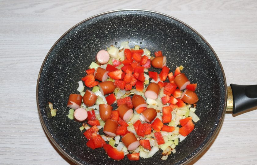 1. Этап. Фасоль по-сербски: К луку и колбаскам добавьте перец, продолжайте обжаривать.