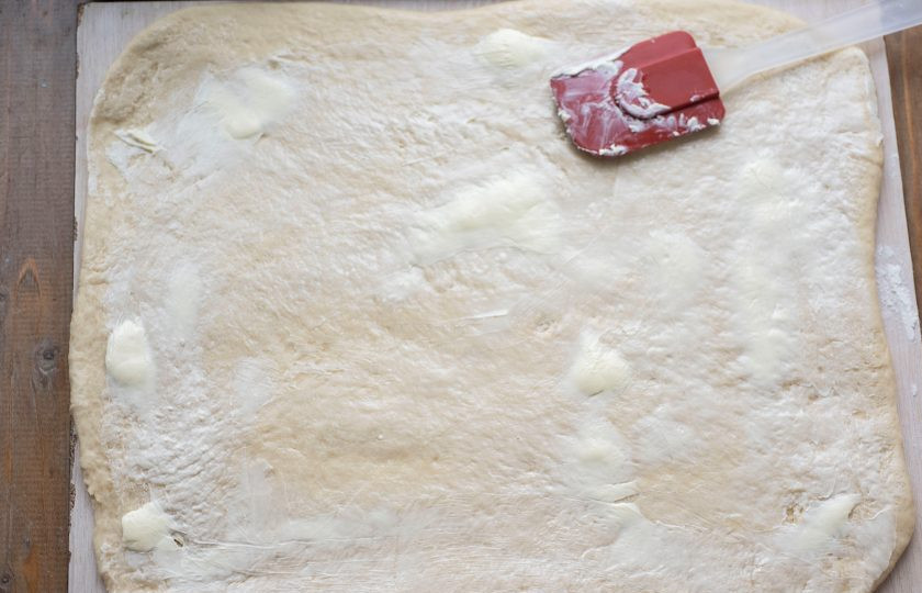 1. Этап. Булочки с яблоком и корицей из дрожжевого теста: Рабочую поверхность припылите мукой, раскатайте тесто скалкой в пласт в форме прямоугольника. Смажьте поверхность мягким сливочным маслом (40 грамм).