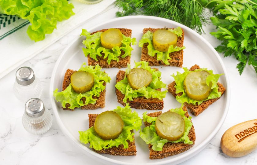 1. Этап. Канапе со шпротой: Промойте листовой салат и нарвите его на полоски. Выложите их на каждый ломтик хлеба, а сверху — кружок соленого огурца.