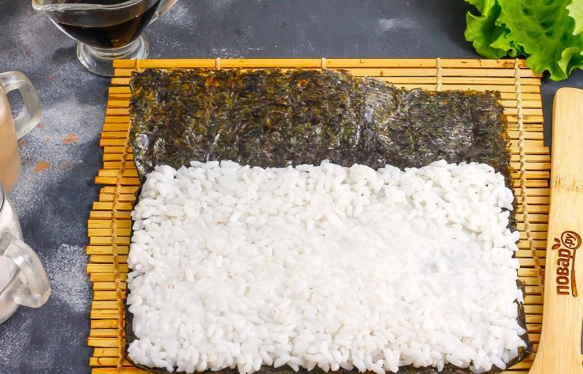 1. Этап. Роллы с курицей и ананасом: Выложите на коврик лист нори, на него выложите часть отварного риса. Смочите руки в воде и распределите рис по водорослям, оставляя один край незаполненным примерно на 2-3 см.