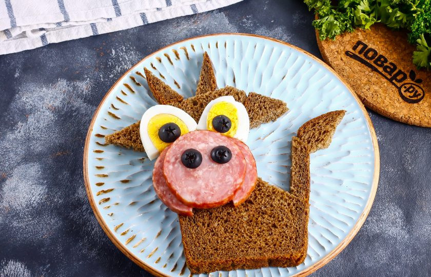 1. Этап. Бутерброд Веселый Бычок: Нарежьте колбасу кружочками и один из них разрежьте пополам. Выложите мордочку быка, на колбасу разложите ноздри из маслин.