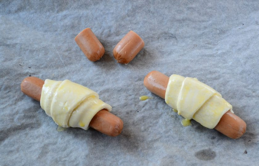 1. Этап. Закуска Крабик: Выложите рогалики на противень, смажьте взбитым яйцом. Одну сосиску разрежьте на 4 равные части. Сделайте в кусочках вертикальные надрезы, примерно до половины. Эти кусочки также положите на противень и запекайте вместе с рогаликами.