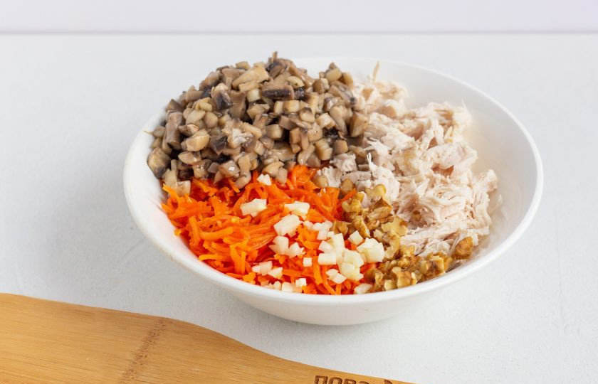 1. Этап. Салат Новогодний подарочек: Шампиньоны мелко нарежьте и обжарьте 2-3 минуты в растительном масле, подсолите. В миске соедините нарезанное кубиками филе, обжаренные шампиньоны, рубленную морковь по-корейски и чеснок, также кусочки орехов.