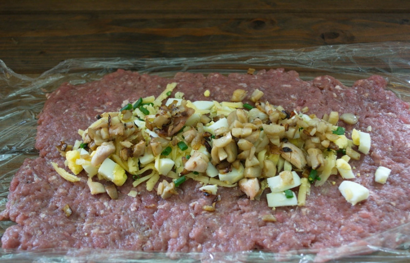 1. Этап. Мясной рулет с грибным соусом: Натрите на терке сыр. Выложите на середину пласта мясного. Отварное яйцо нарезать кубиком и выложить на сыр. Распределить. Шампиньоны мелко нарезать. На сковороде обжарьте шампиньоны с луком. Выложите сверху на сыр.