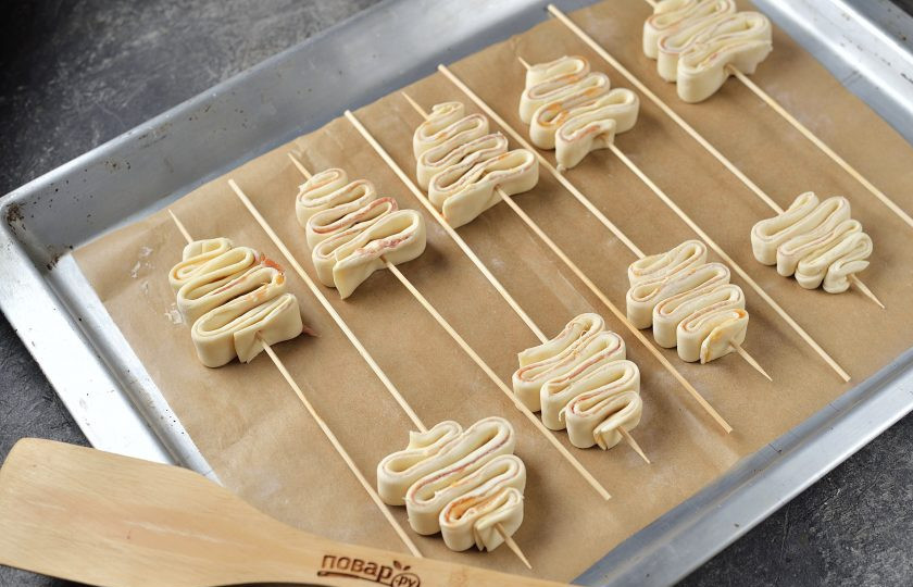 1. Этап. Канапе «Ёлочка» из слоеного теста: Далее тесто нужно нанизать на бамбуковые шпажки, придав ему форму елочек. При желании заготовки можно смазать взбитым яйцом. Отправьте противень в предварительно разогретую духовку в режиме 