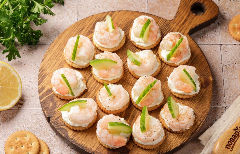1. Этап. Канапе с креветкой на крекере: Огурец нарежьте кружочками, а затем каждый кружок разрежьте на 2 или 4 части. Украсьте канапе огурцом и любой свежей зеленью.