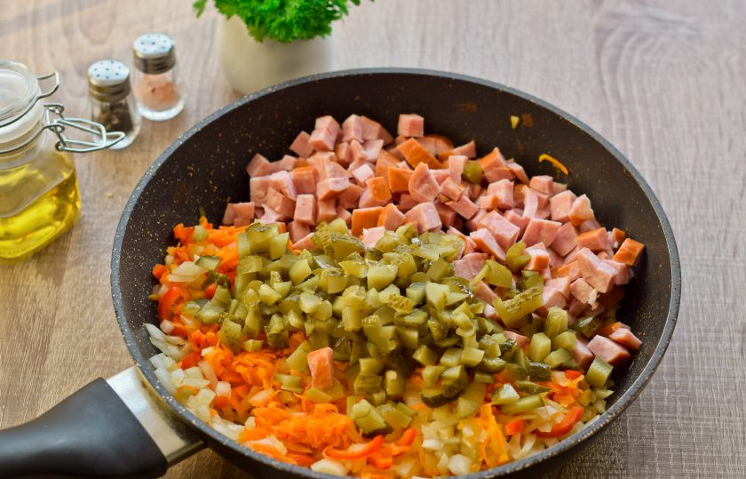 1. Этап. Рассольник с копченой колбасой и перловкой: В сковороде разогрейте растительное масло, выложите сначала лук, морковь, перец, жарьте в течение 10 минут, помешивая. После нарежьте копченую колбасу кубиками и добавьте в сковороду. Также нарежьте небольшими кусочкам корнишоны, добавьте в сковороду.