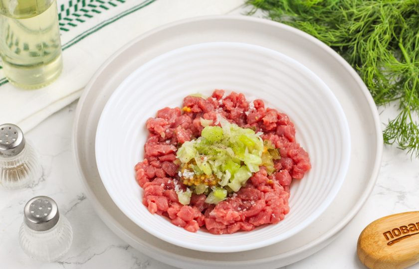 1. Этап. Тартар из говядины классический: Промойте и измельчите лук-порей или красный лук, добавляя нарезку в емкость вместе с маслом. Посолите все и поперчите, тщательно перемешайте и оставьте на 30 минут в холодильнике. По желанию можно добавить соус Табаско или вустерский соус. Они придадут блюду легкую пикантность.