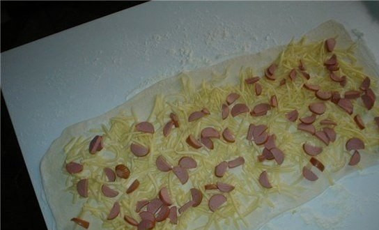 1. Этап. Сырная улитка: Возьмите тесто и разрежьте на полоски. Ширина каждой полоски примерно 7 см, а длина примерно 20 см. С помощью скалки раскатайте полоски теста. Не давите сильно, это важно. Тонкие полоски делать не нужно, они быстро порвутся. Затем по полоске равномерно распределяем начинку: сыр с колбасой.